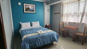 Quarto com cama, uma mesinha, duas cadeiras, um quadro na parede e janela com cortinas.