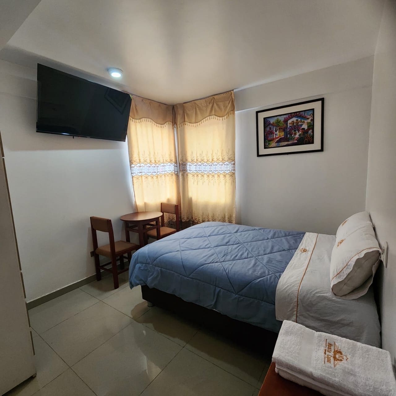 Een slaapkamer met een bed, een bureau en een stoel. Er is een televisie aan de muur en een schilderij.