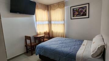 Een slaapkamer met een bed, een bureau en een stoel. Er is een televisie aan de muur en een schilderij.