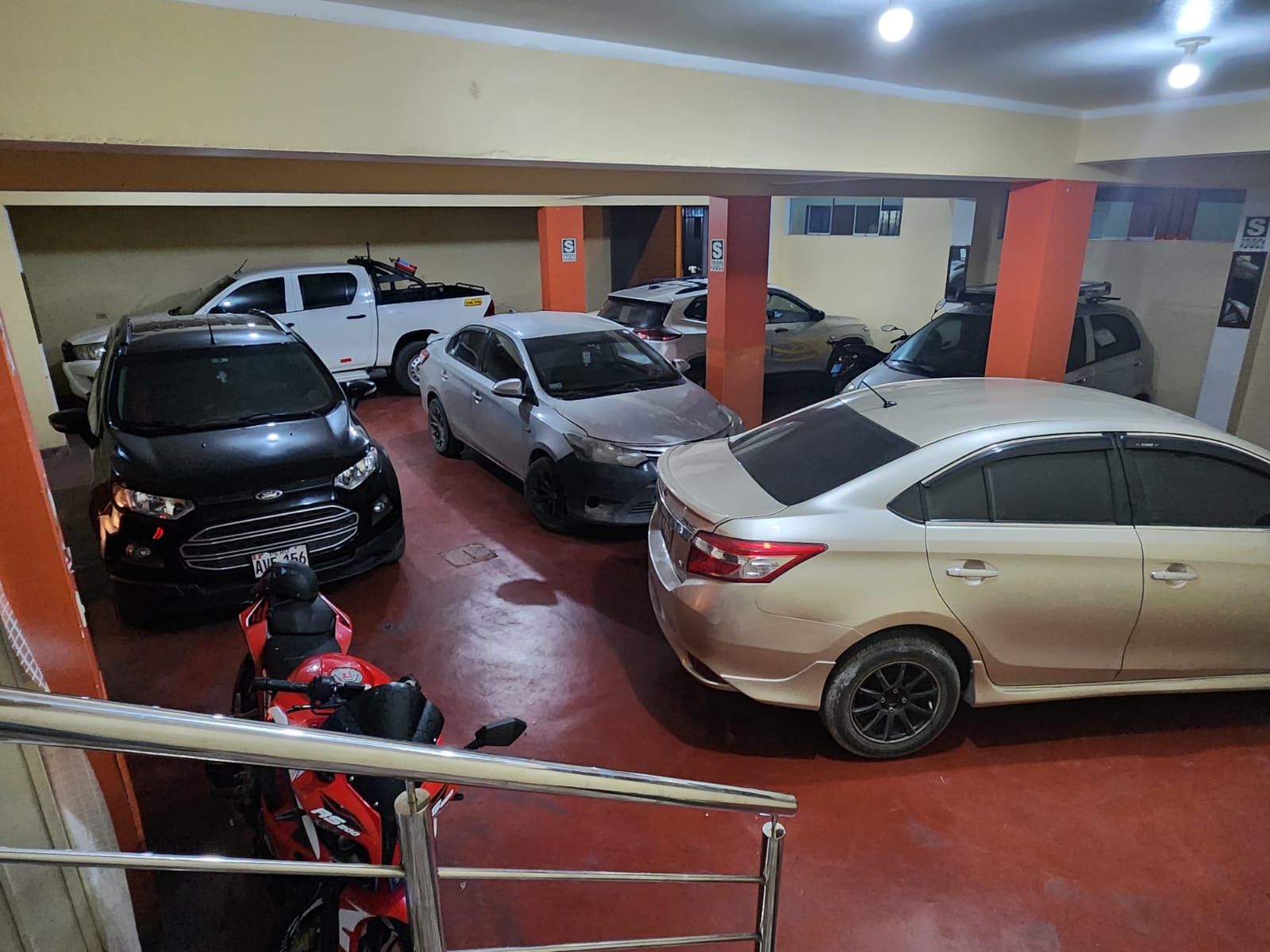 A parking garage with multiple cars and a motorcycle, featuring concrete pillars and a red floor.