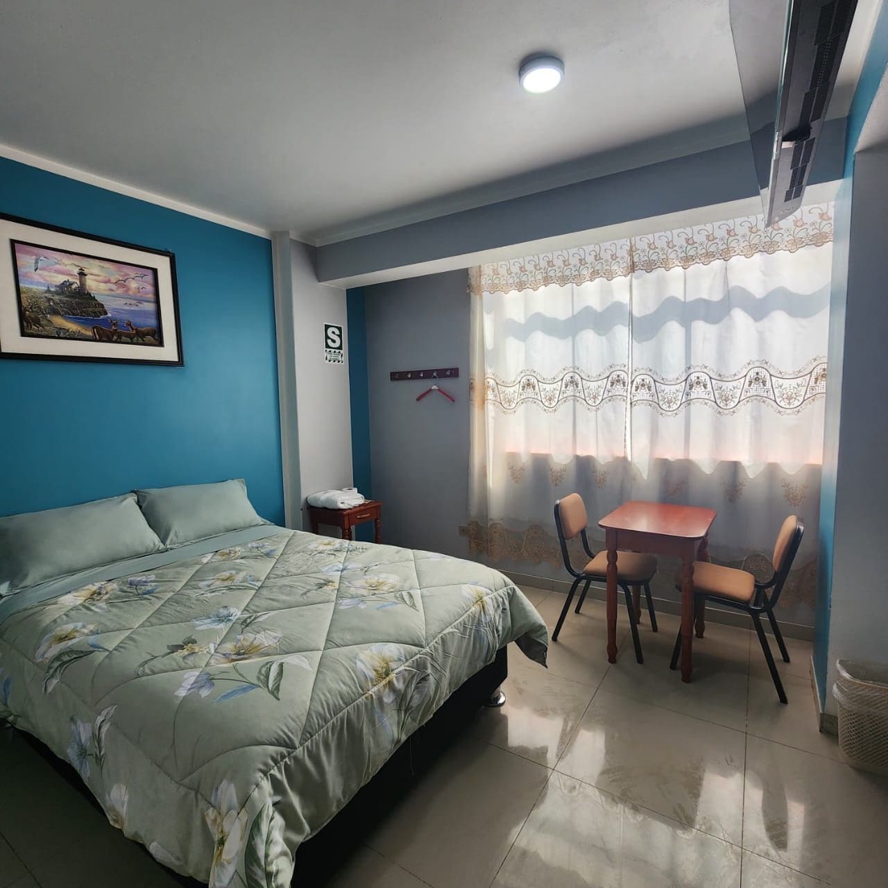 Quarto com cama, mesa com cadeiras, um quadro na parede e janela com cortinas.