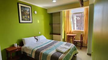 A hotel room with a bed, a bedside table, a small table with chairs, a window with curtains, and a wall-mounted light fixture.