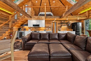 A modern log cabin interior with a large leather sectional sofa, a kitchen with white cabinets, and a wooden staircase.