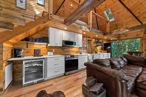 A cozy cabin interior with a kitchen, a brown leather sofa, and a large window.