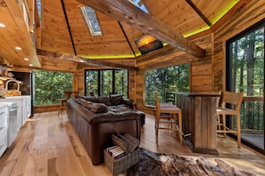 A cabin interior with a sofa, dining table, chairs, and a view of the forest.