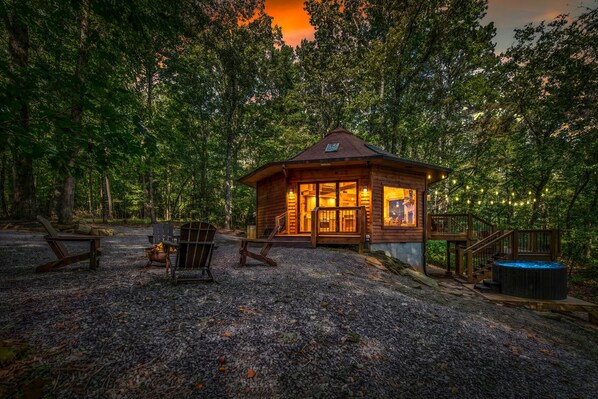 A wooden cabin with a porch, surrounded by trees, and equipped with outdoor seating and a fire pit.
