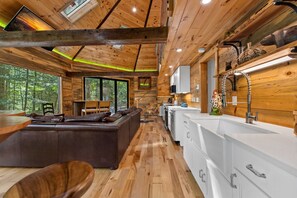 A cabin interior with a wooden ceiling, a kitchen area with white cabinets, and a seating area with a brown leather sofa.