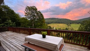 Una terraza de madera con una mesa y un banco, con vistas a un paisaje verde con colinas y un árbol.