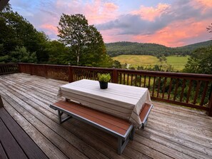 Une terrasse en bois avec une table et un banc, donnant sur un paysage verdoyant avec des collines et un arbre.