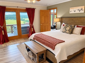 Une chambre avec un grand lit, une tête de lit en bois, un banc, une vue sur l’extérieur grâce à une porte coulissante, et un tableau accroché au mur.