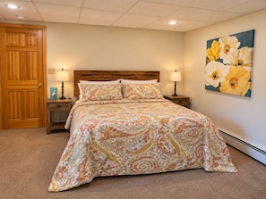A bedroom with a wooden door, a bed with patterned bedding, two bedside tables with lamps, and a floral painting on the wall.