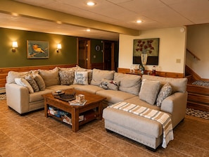 A living room with a sectional sofa, a coffee table, and a bench. There is a wall clock and a painting above the sofa.