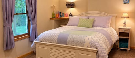 A bedroom with a bed, a nightstand, a lamp, a shelf with books, and a window with curtains.