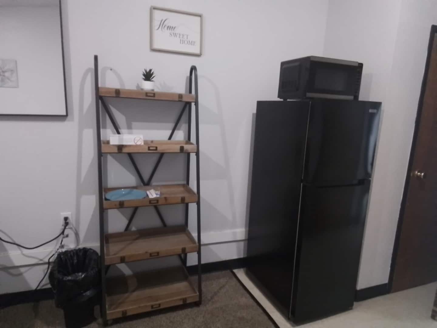 A kitchen with a black refrigerator, a wooden shelf unit, and a trash bin.