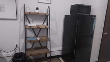 A kitchen with a black refrigerator, a wooden shelf unit, and a trash bin.