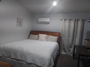 A bedroom with a bed, a bedside table, a lamp, a wall-mounted air conditioner, and a window with curtains. - The Garden Inn (Sinton)