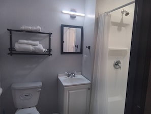 A bathroom with a toilet, sink, shower, towel rack, and a mirror. - The Garden Inn (Sinton)