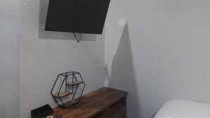Et værelse med et monteret fjernsyn på væggen, en træbænk med en geometrisk hylde og en seng med hvide sengetæpper.