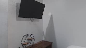 A room with a mounted TV on the wall, a wooden bench with a geometric shelf, and a bed with white bedding.