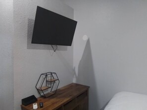 A room with a mounted TV on the wall, a wooden bench with a geometric shelf, and a bed with white bedding. - The Garden Inn (Sinton)