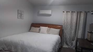 A bedroom with a bed, a bedside table, a lamp, a wall-mounted air conditioner, and a window with curtains.