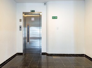 Ein Flur mit einer Metall-Lift-Tür, einem grünen Fluchtzeichen und einem an der Wand befestigten Lichtschalter.