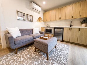 A modern living room with a grey sofa, a matching ottoman, a patterned rug, a wall-mounted air conditioner, and wooden cabinets. - Modern 3-bedroom Capitol Hill Home with Full Kitchen (Seattle)