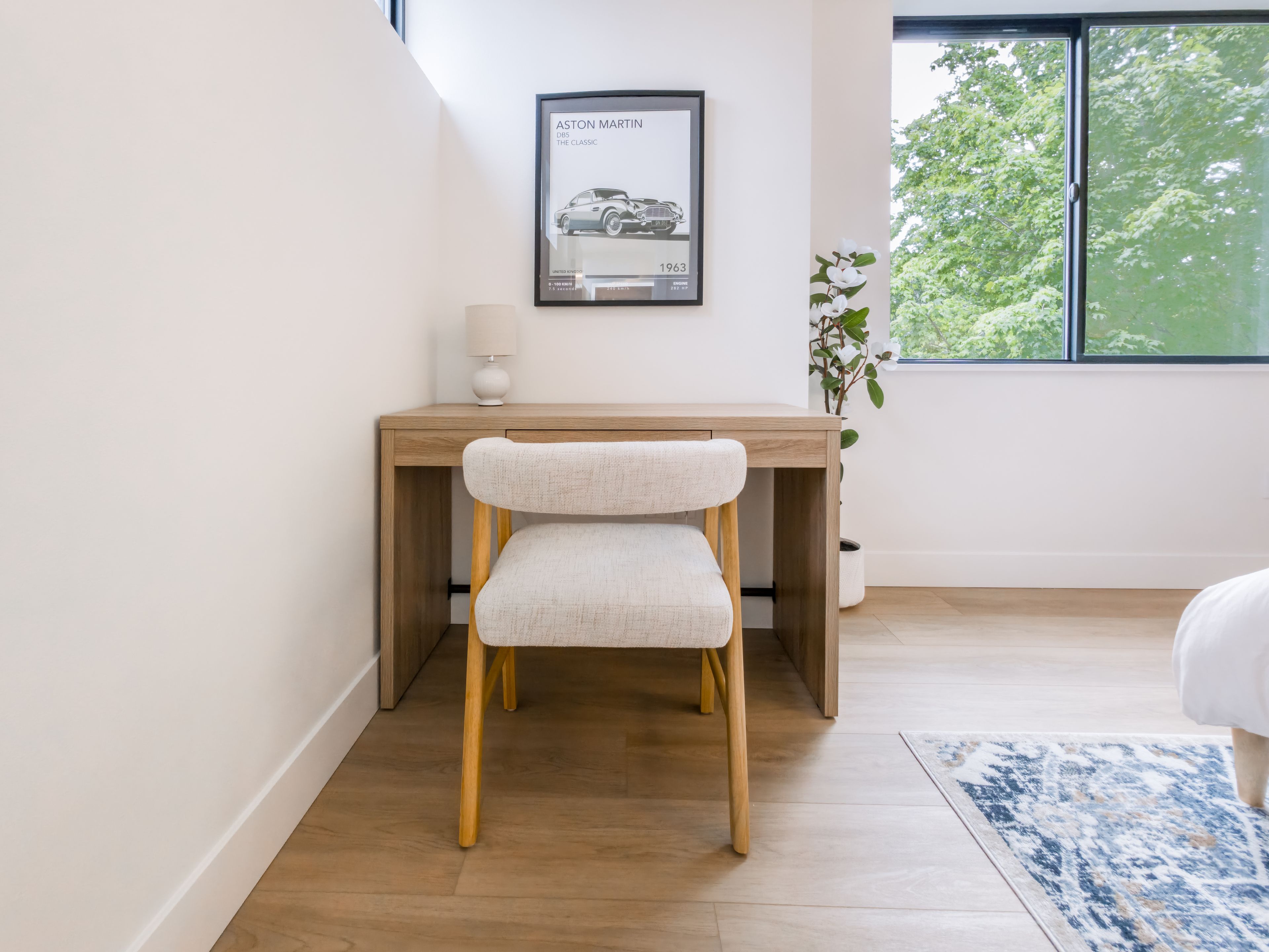 Een moderne kantoorruimte met een houten bureau, een witte fauteuil, een lijst met een afbeelding aan de muur, en een raam met uitzicht op bomen.