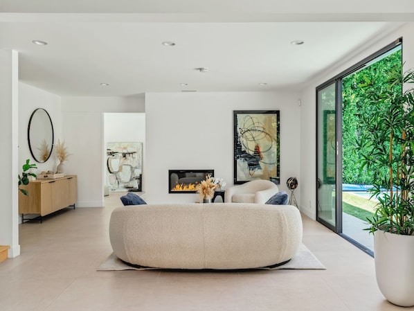 A modern living room with a large bean bag, a fireplace, and a large window with a view of greenery. - Casa Leona (Encino)