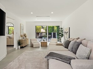 A modern living room with a beige sofa, a coffee table, and a large window offering a view of the outdoors. - Casa Leona (Encino)