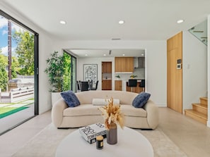 A modern living room with a beige sofa, a coffee table, and a book titled "TOM FORD CO." A plant is on the coffee table, and the room has a large window. - Casa Leona (Encino)