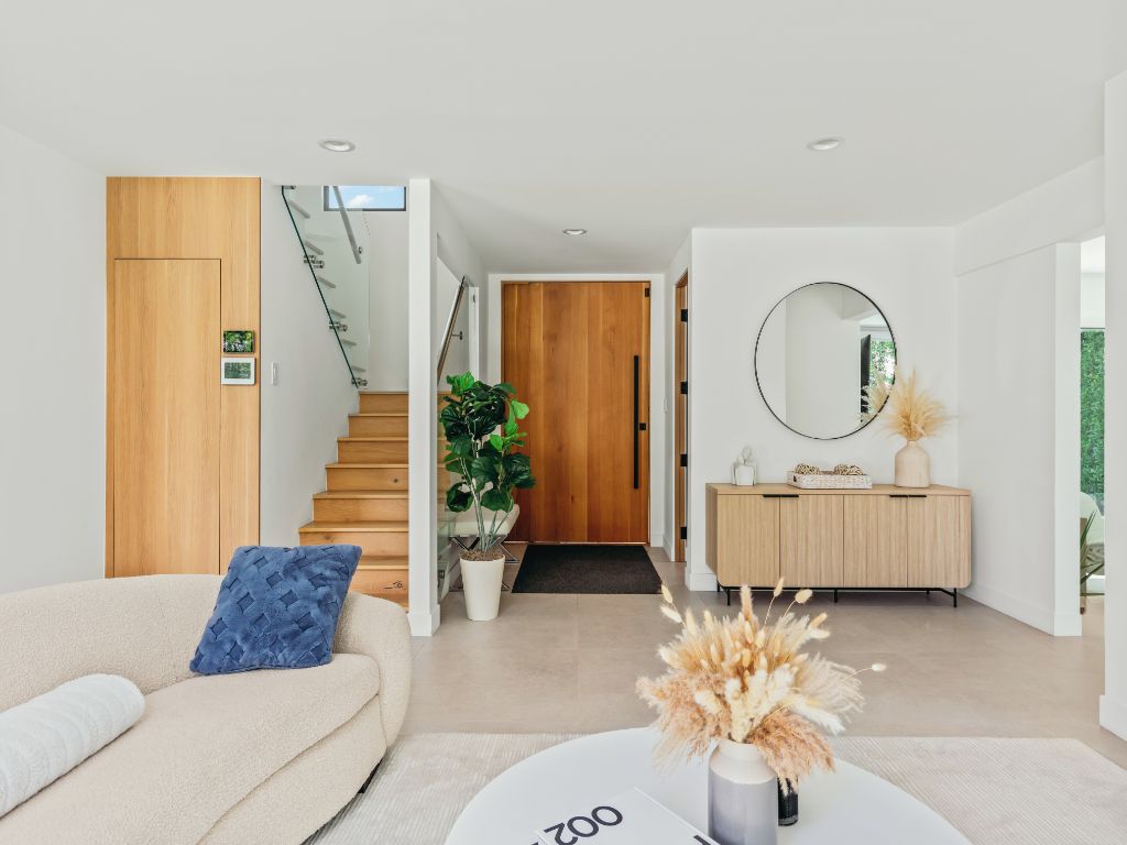 Una sala moderna con un sofá, una mesa de centro con un jarrón, una escalera, una puerta de madera y un espejo en la pared.