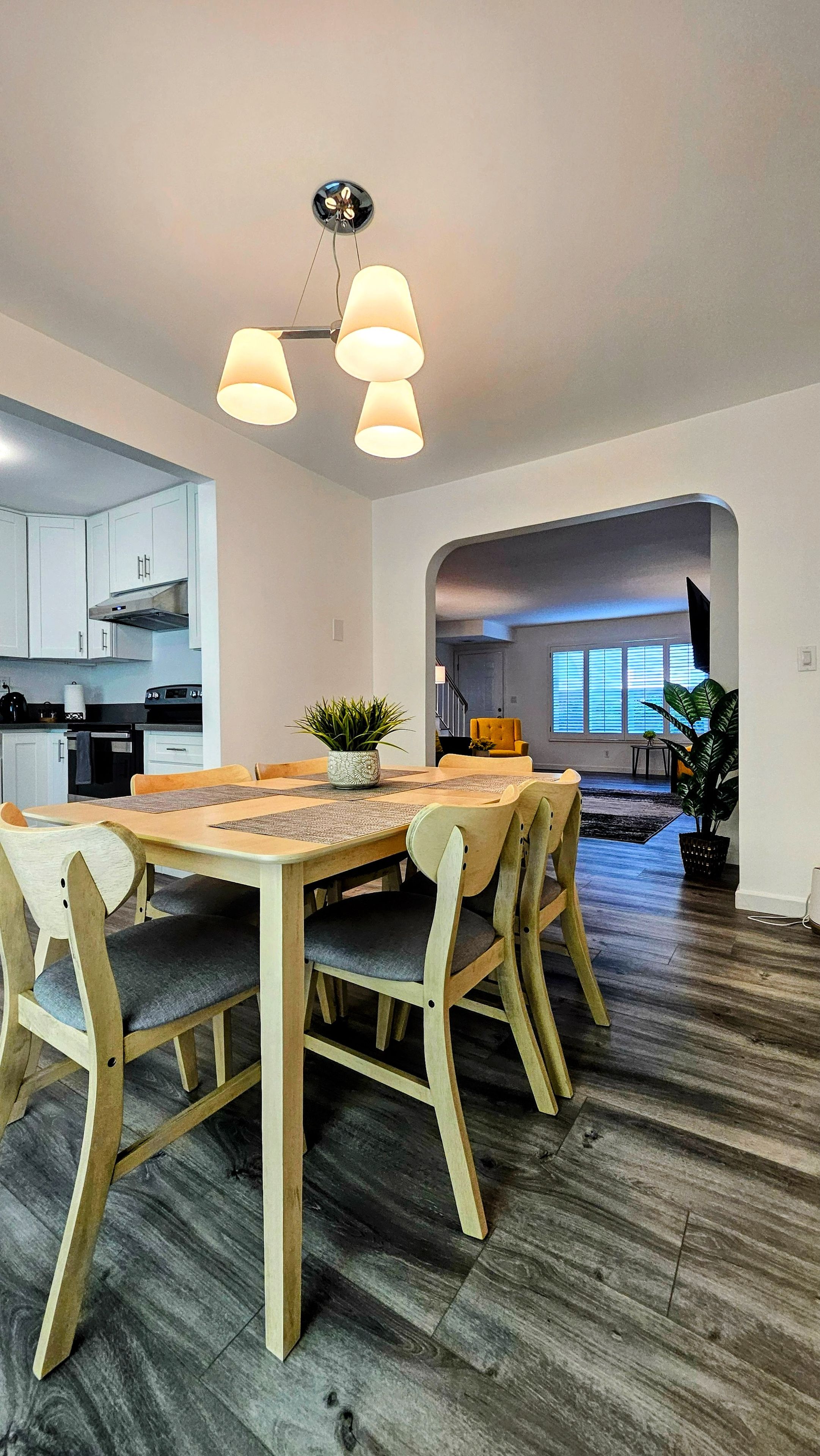 Eine moderne Küche mit weißen Schränken, ein Essbereich mit Holztisch und -stühlen sowie ein Wohnzimmer mit Sofa und großem Fenster.