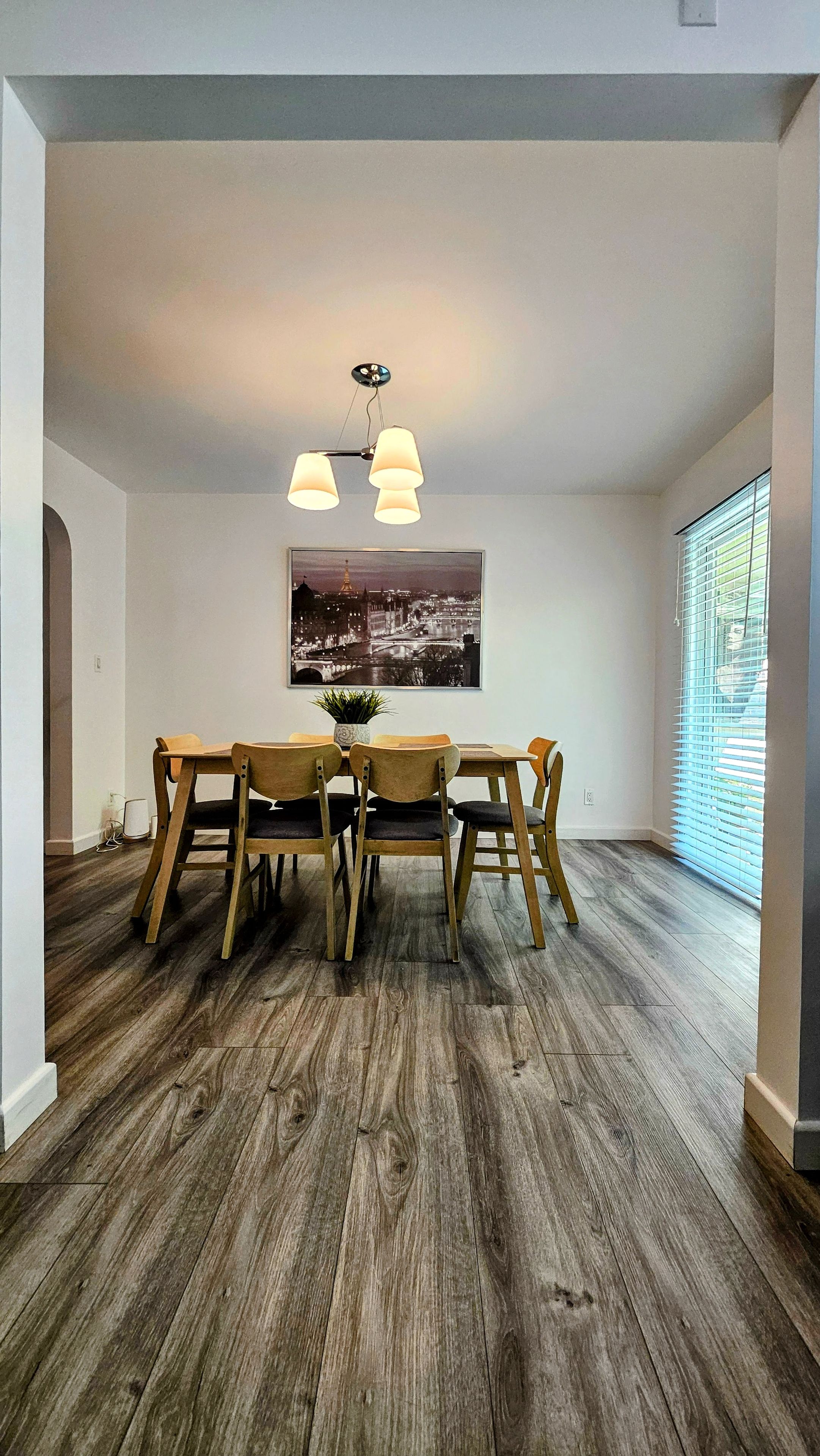 A dining area with a wooden table and chairs, a ceiling light fixture, and a cityscape painting on the wall.