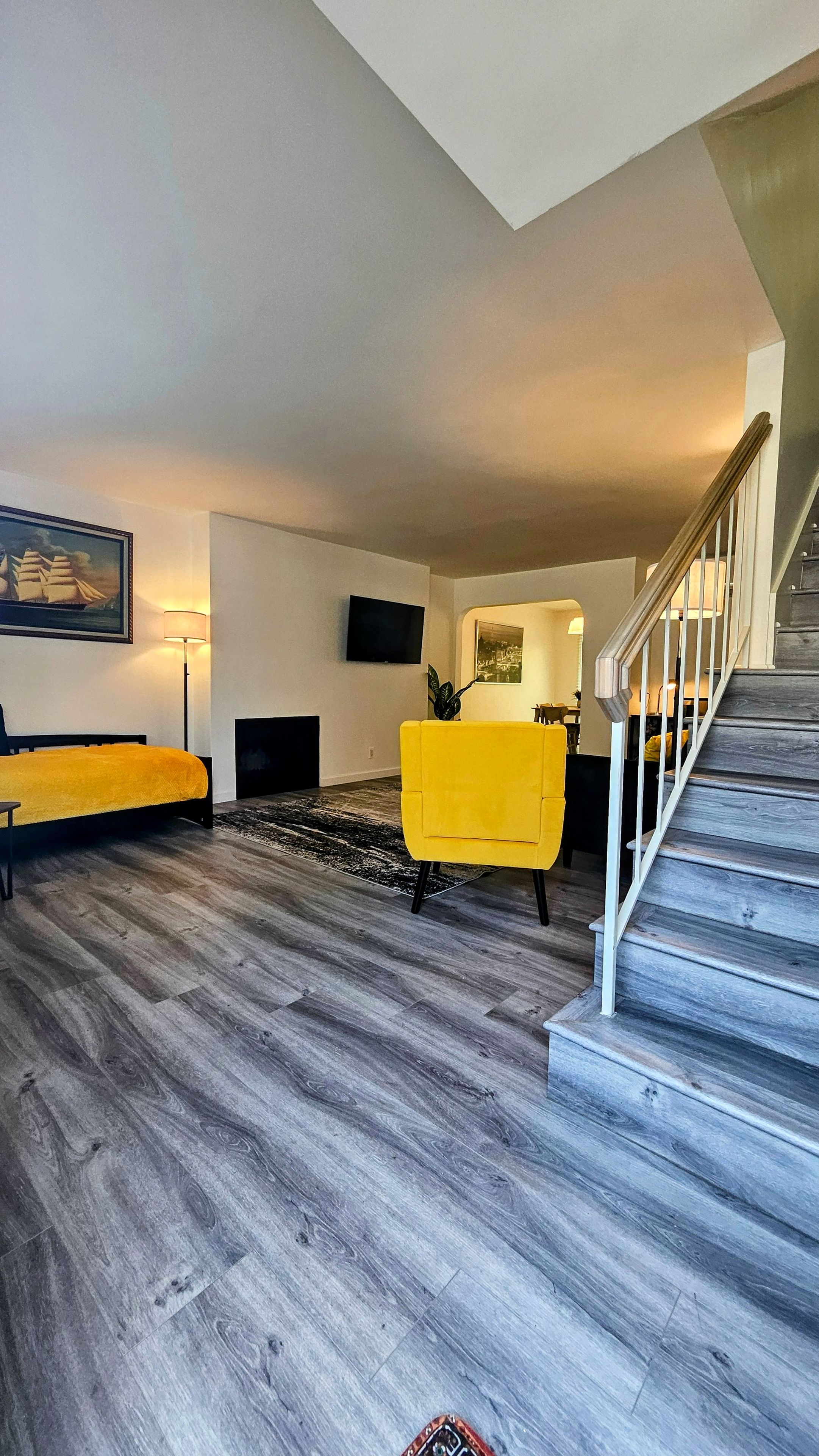A modern living room with a sofa, a yellow armchair, a flat-screen TV mounted on the wall, and a staircase leading to another level.