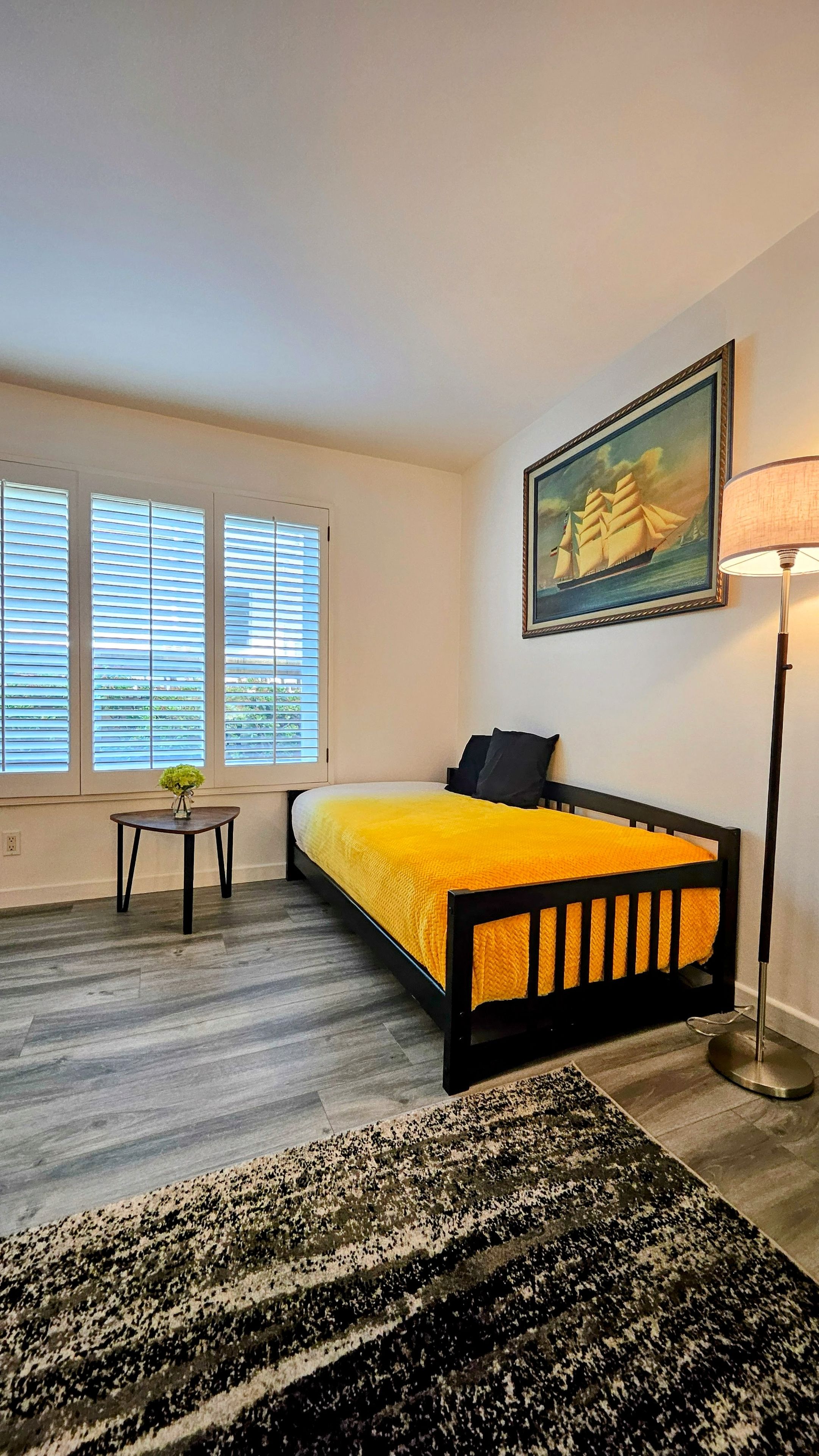 Ein Schlafzimmer mit Bett, einem Gemälde, einer Lampe und einem Fenster mit Jalousien.