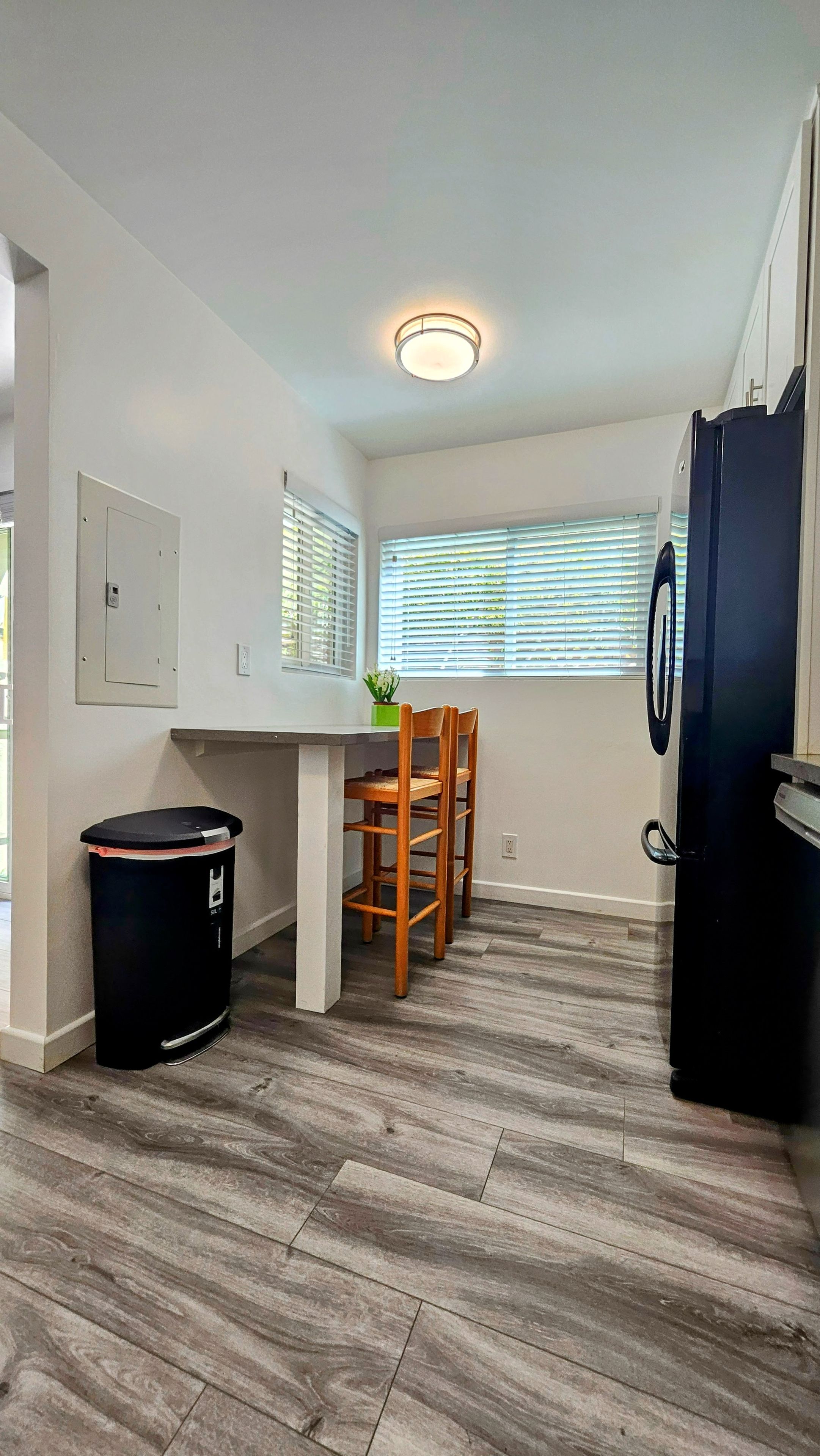 A compact kitchen with a dining table, chairs, a black trash bin, a refrigerator, and a window with blinds.