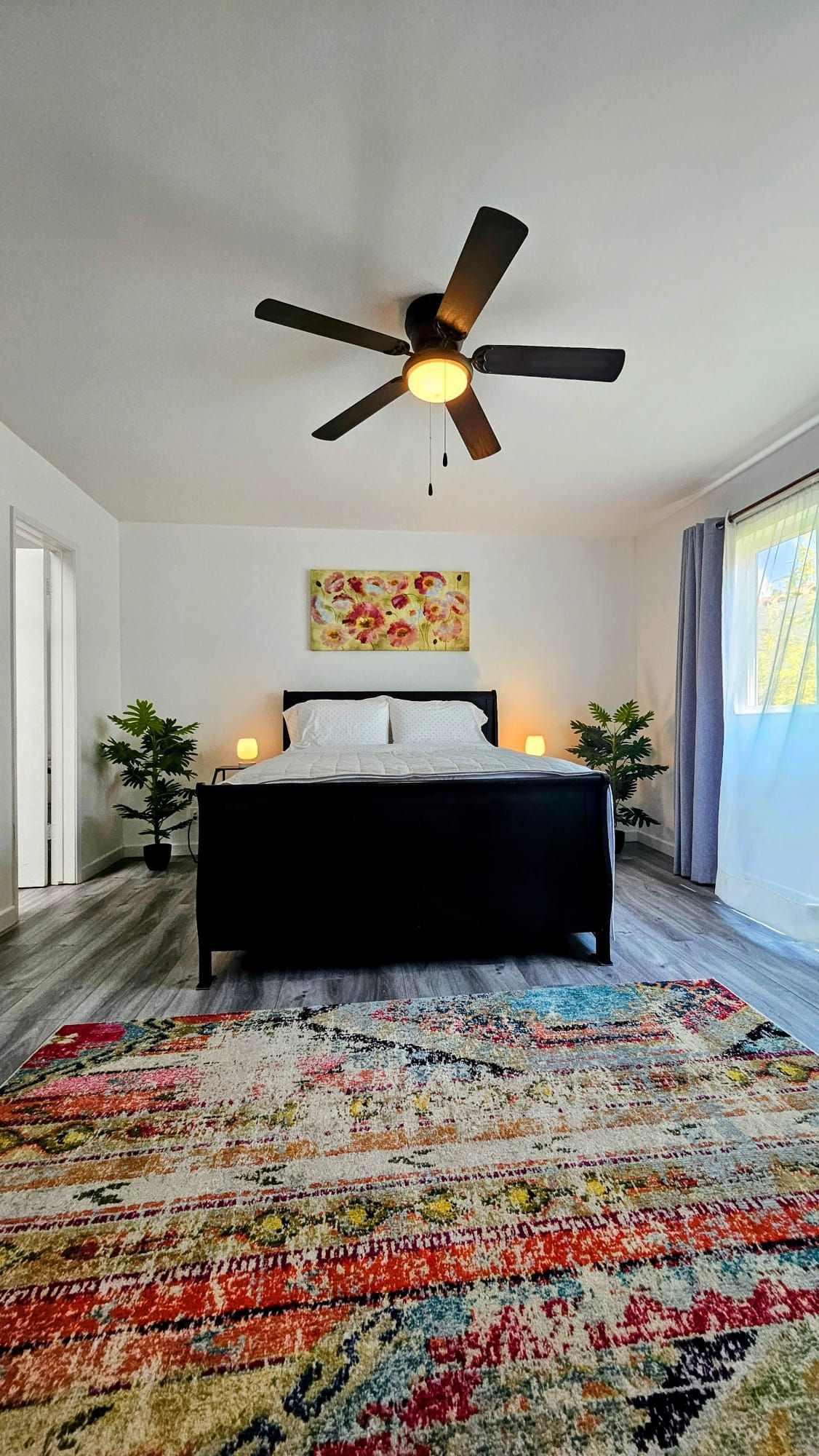 A bedroom with a bed, a ceiling fan, a painting, and a large rug.