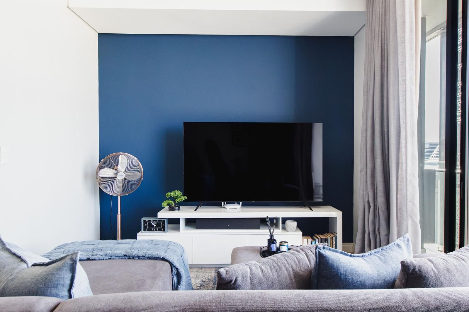 A modern living room with a flat-screen TV on a white stand, a standing fan, and a sofa with cushions.