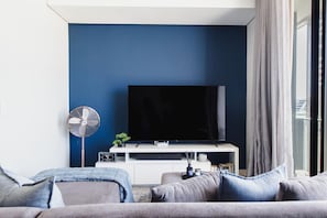 Un saló modern amb un televisor de pantalla plana sobre un suport blanc, un ventilador de peu i un sofà amb coixins.