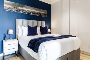 A modern hotel room with a large bed, a headboard, a nightstand, a lamp, and a cityscape painting on the wall.
