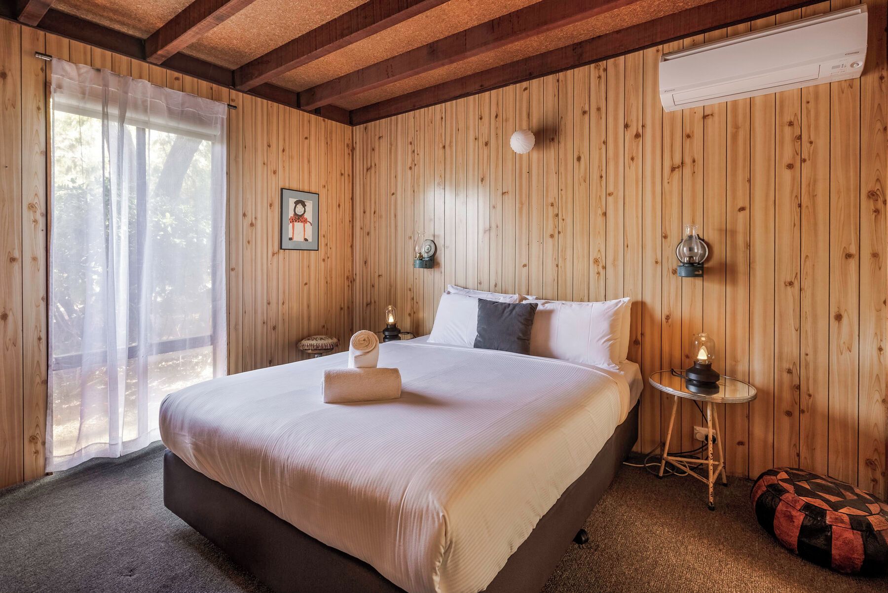 A bedroom with a wooden paneled wall, a bed with white bedding, a bedside table with a lamp, and a window with sheer curtains.