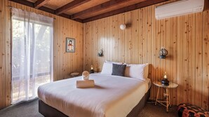 A bedroom with a large bed, a bedside table with a lamp, a wall-mounted air conditioner, and a window with sheer curtains. - New: Quitch Cottage - steps from the beach (Quindalup)