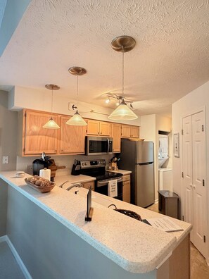 A kitchen with wooden cabinets, a stainless steel refrigerator, a microwave, and a stove. There are pendant lights hanging from the ceiling. - Coastal 2B condo minutes from the beach & attractions with pool and golf access! (Longs)