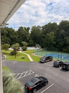 Un aparcament amb una pista de tennis, un edifici i una zona boscosa.