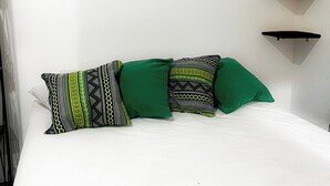 Uma cama com colchão branco, três almofadas com padrões geométricos e três prateleiras pretas fixadas na parede.