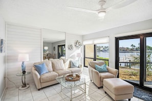 A modern living room with a glass coffee table, two sofas, a lamp, and a view of a body of water through large windows.