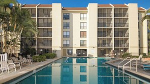 Uma área de piscina com piscina azul translúcida, rodeada por espreguiçadeiras e um edifício com várias andares e varandas.