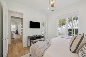 A bedroom with a large bed, a desk with a flat-screen TV, a window with a view of a house, and a door leading to a bathroom. - Seagrove: Private Heated Pool, Casita Incl, Bikes, 3 min walk to Beach (Seagrove Beach)
