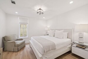 Un dormitorio con una cama grande, una mesita de noche con lámpara, una silla y una ventana con persianas.